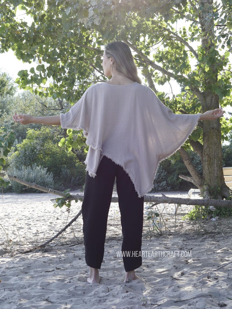 May include: A woman wearing a light brown, loose-fitting, asymmetrical top with a frayed hem and black pants. The top has a wide, flowing silhouette. The woman is standing on a sandy beach with trees in the background. The text "www.hearthcraft.com" is visible at the bottom of the image.