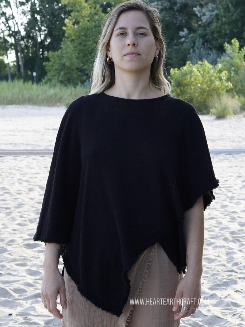 May include: A black cotton poncho with a frayed edge, worn by a person with long blonde hair. The poncho is draped over a brown skirt. The person is standing on a sandy beach.