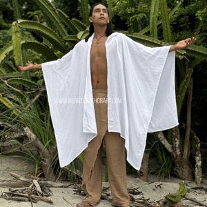 Pu&ograve; includere: Un poncho bianco in garza di cotone con cappuccio e un design lungo e fluido. Il poncho &egrave; indossato sopra pantaloni marroni e il modello &egrave; in piedi in un ambiente naturale.