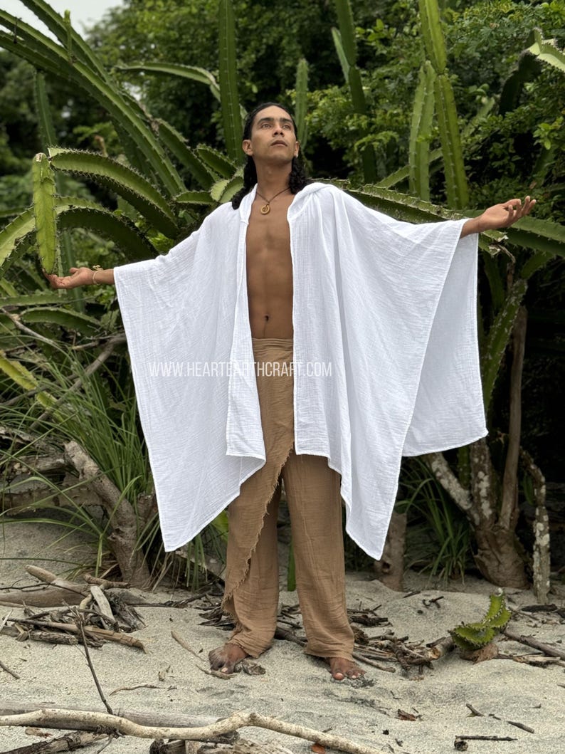 Pu&ograve; includere: Un poncho bianco in garza di cotone con cappuccio e un design lungo e fluido. Il poncho &egrave; indossato sopra pantaloni marroni e il modello &egrave; in piedi in un ambiente naturale.