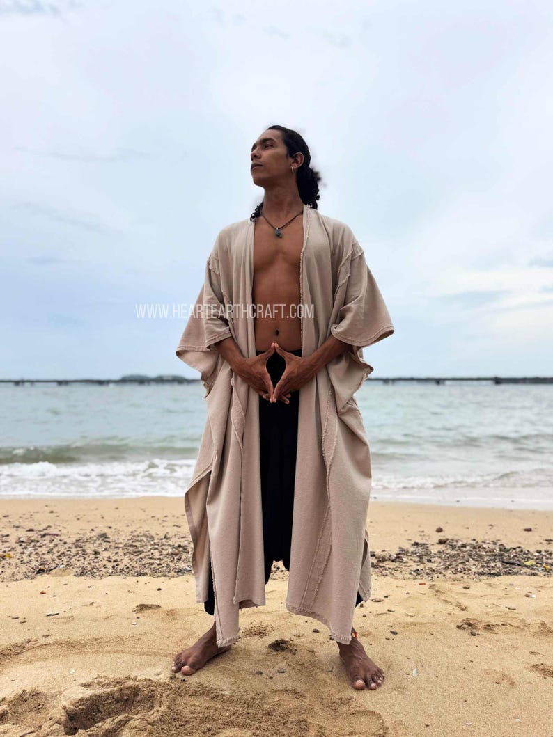 Pu&ograve; includere: Un accappatoio beige in stile kimono con maniche larghe e orlo sfrangiato. L'accappatoio &egrave; indossato sopra pantaloni neri. L'immagine &egrave; scattata su una spiaggia sabbiosa con l'oceano sullo sfondo. Il sito web www.heartearthcraft.com &egrave; visibile.