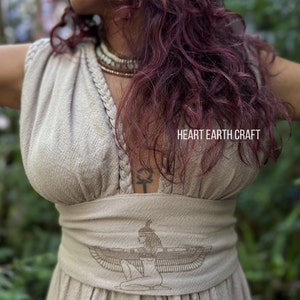 May include: A beige linen dress with a V-neckline and a braided detail. The dress has a wide waistband with an embroidered image of a winged goddess. The text "HEART EARTH CRAFT" is visible on the dress.