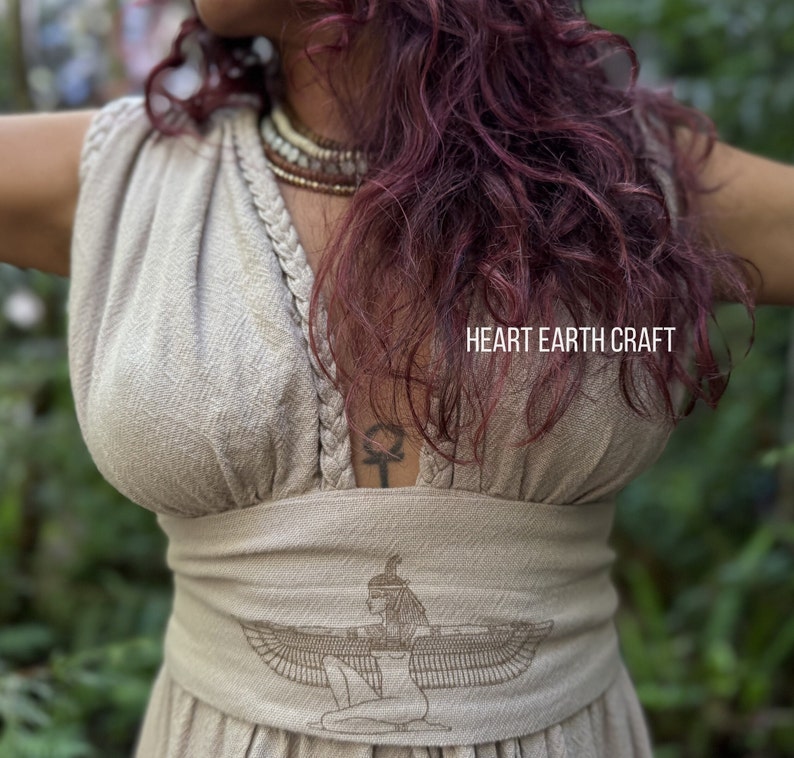 May include: A beige linen dress with a V-neckline and a braided detail. The dress has a wide waistband with an embroidered image of a winged goddess. The text "HEART EARTH CRAFT" is visible on the dress.