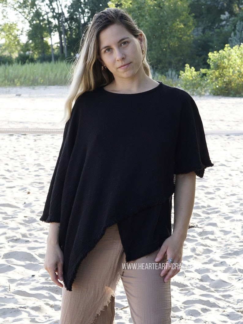 May include: A black, asymmetrical, short-sleeved top with a raw edge hem. The top is worn with brown pants and the model is standing on a sandy beach.