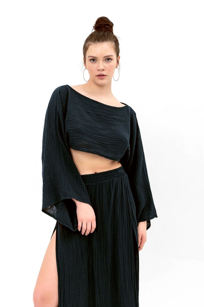 May include: A black two-piece outfit with a cropped top and a long skirt with a side slit. The top has a wide, boat neckline and long sleeves. The skirt has a wide waistband and a flowing silhouette.