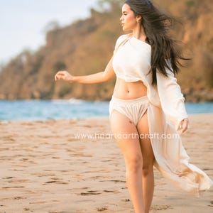 May include: A cream-colored two-piece set consisting of a cropped, one-shoulder top with long sleeves and matching high-waisted shorts with an attached flowing side panel. The fabric has a textured finish. The text www.heartearthcraft.com is visible.