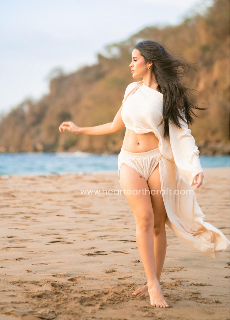 May include: A cream-colored two-piece set consisting of a cropped, one-shoulder top with long sleeves and matching high-waisted shorts with an attached flowing side panel. The fabric has a textured finish. The text www.heartearthcraft.com is visible.