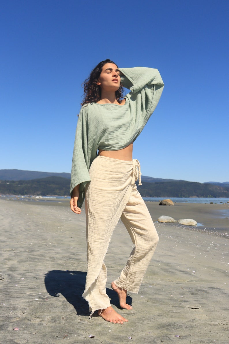 May include: A woman wearing a light green, loose-fitting, long-sleeved top and cream-colored pants with a wrap-style waist. She is standing on a sandy beach with a blue sky in the background.
