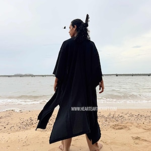 Pu&ograve; includere: Una persona indossa un lungo abito nero in stile kimono su una spiaggia sabbiosa, di fronte all'oceano. L'abito ha maniche larghe e uno spacco frontale. Un uccello vola nel cielo. Il sito web www.heartearthcraft.com &egrave; visibile.
