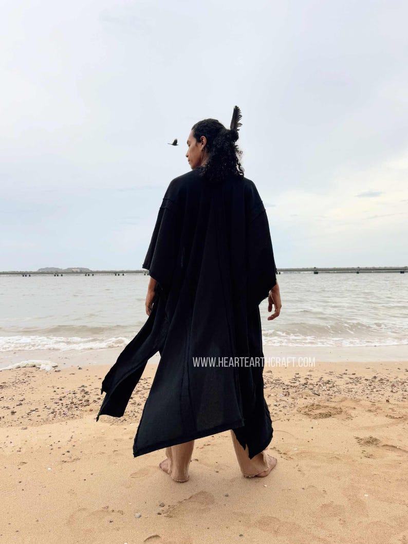 Pu&ograve; includere: Una persona indossa un lungo abito nero in stile kimono su una spiaggia sabbiosa, di fronte all'oceano. L'abito ha maniche larghe e uno spacco frontale. Un uccello vola nel cielo. Il sito web www.heartearthcraft.com &egrave; visibile.