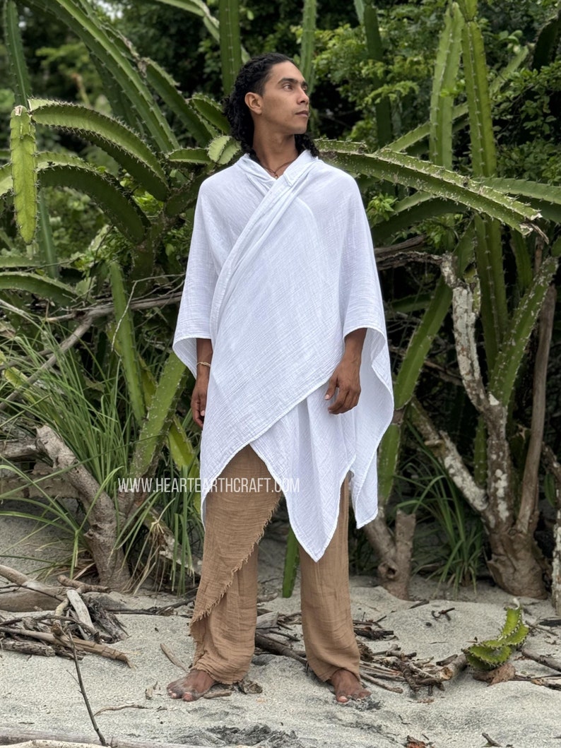 Pu&ograve; includere: Un poncho bianco in cotone con cappuccio e un design lungo e fluido. Il poncho &egrave; indossato sopra pantaloni marroni con orlo sfrangiato. La persona &egrave; in piedi davanti a una pianta di cactus.