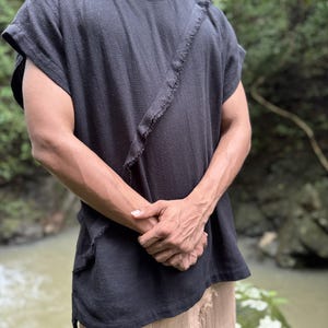 May include: Black short-sleeved shirt with a raw, diagonal seam detail. The shirt has a relaxed fit and a textured appearance. The model is wearing tan pants. The background is a natural outdoor setting with a river.