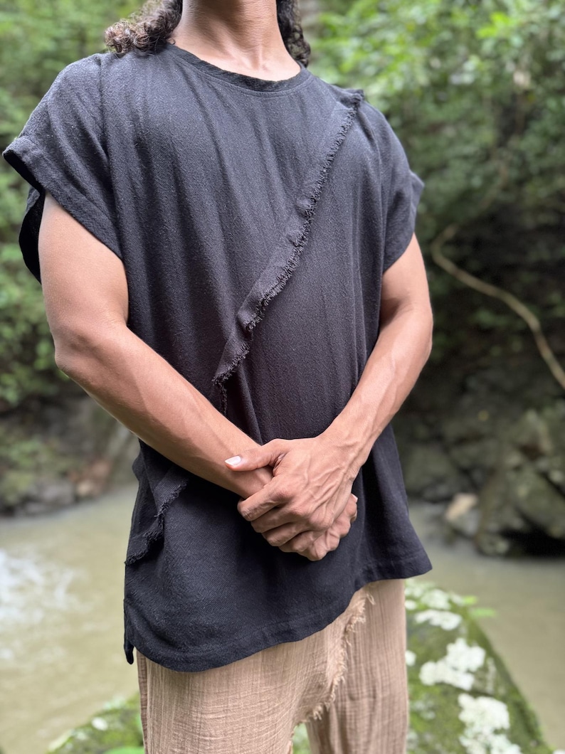 May include: Black short-sleeved shirt with a raw, diagonal seam detail. The shirt has a relaxed fit and a textured appearance. The model is wearing tan pants. The background is a natural outdoor setting with a river.