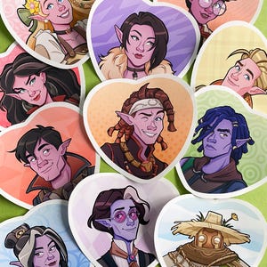 May include: A collection of heart-shaped stickers featuring fantasy character portraits. Each sticker showcases a different character with unique features, including pointed ears, various hairstyles, and diverse skin tones. The stickers are arranged on a green surface.