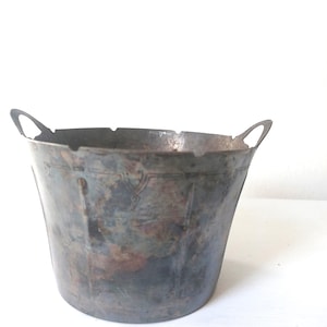 May include: A vintage metal bucket with two arched handles. The bucket has a weathered, tarnished appearance with shades of gray, brown, and blue. The bucket's shape is slightly tapered, and it appears to be made of a sturdy metal.