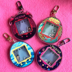 Könnte beinhalten: Vier Acryl-Schlüsselanhänger mit dem Text "FALL OUT BOY" in verschiedenen Farben und Designs. Jeder Schlüsselanhänger hat einen kleinen Bildschirm mit Miniaturfiguren. Die Schlüsselanhänger haben gold- oder silberfarbene Verschlüsse und sind auf einem rosa Hintergrund platziert.