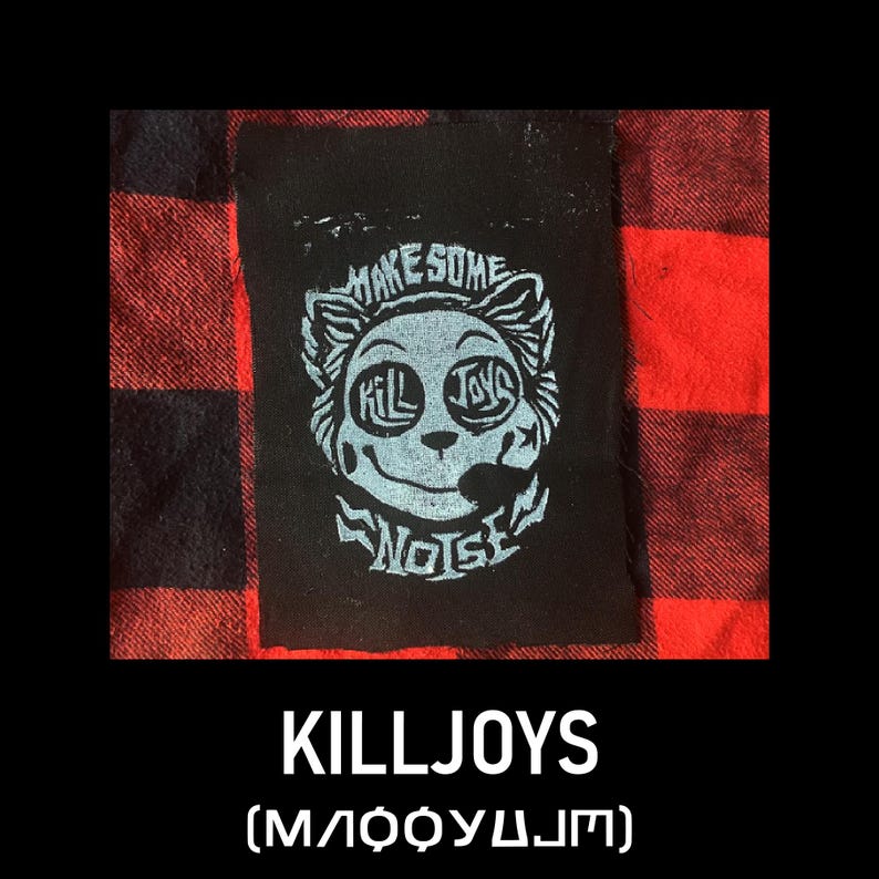 May include: Black patch featuring a light blue cartoon face design with the words "MAKE SOME NOISE" and "KILLJOYS". The patch is set against a red and black plaid background. The word "KILLJOYS" is printed below the patch.