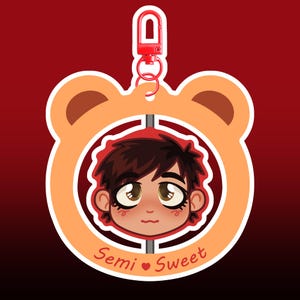Puede incluir: Un llavero de dibujos animados con un personaje de pelo castaño dentro de un marco con forma de oso naranja. El llavero tiene un cierre y un anillo rojos. Las palabras "Semi Sweet" están escritas en cursiva debajo de la cara del personaje.