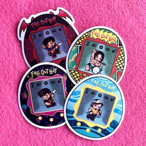Puede incluir: Cuatro pegatinas coloridas con ilustraciones de dibujos animados de miembros de la banda tocando instrumentos. Cada pegatina tiene el texto "Fall Out Boy" y un diseño que se asemeja a un juego portátil retro. Las pegatinas están sobre una superficie rosa.
