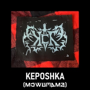 May include: Black fabric patch with a light blue graphic design and the word "KEPOSHKA" in white text. The patch is set against a red and black plaid background. Below the word "KEPOSHKA" is the text "(MƆWUM&Delta;MZ)".
