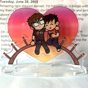 Könnte beinhalten: Ein klarer Acrylständer mit einem herzförmigen Design, das zwei Cartoonfiguren in einer Umarmung zeigt. Das Herz hat einen Farbverlauf aus Rosa, Orange und Gelb. Die Figuren sitzen auf einem braunen Ast. Im Hintergrund Text: "Tuesday, June 28, 2005".