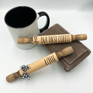 May include: Two wooden rolling pins with black text. One says "But First Coffee" and the other says "Latte". The rolling pin with the "Latte" text has a black and white checkered ribbon tied around it.