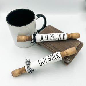 May include: Two white painted rolling pins with black lettering. One rolling pin says "JUST BREW IT" and the other says "GOT MILK". Both rolling pins have a black and white checkered ribbon tied around them.