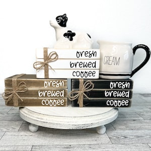 May include: Three sets of stacked wooden blocks with the text "fresh brewed coffee" on each. The blocks are painted white, gold, and black. A white ceramic creamer with the word "CREAM" on it is sitting on top of the blocks. A white ceramic cow figurine is sitting on the blocks to the left of the creamer.