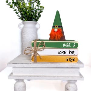 May include: A white tiered stand with three stacked books. The books are painted green, white, and gold with the text "Just a wee bit Irish". A green and orange gnome figurine sits on top of the books.
