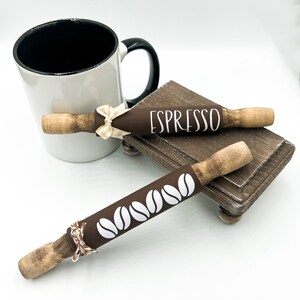 May include: Two brown wooden rolling pins with white designs. One rolling pin has the word "ESPRESSO" written on it. The other rolling pin has a design of coffee beans. The rolling pins are sitting on a small wooden stand.