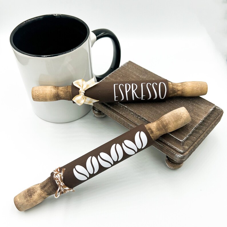 May include: Two brown wooden rolling pins with white designs. One rolling pin has the word "ESPRESSO" written on it. The other rolling pin has a design of coffee beans. The rolling pins are sitting on a small wooden stand.