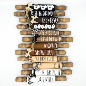 May include: A set of ten wooden rolling pins with various coffee-themed sayings painted on them. The sayings include "Rise & Grind", "Espresso", "Cappuccino", "Fresh Brewed", "Mocha", "Brew-tiful", "But First Coffee", "Latte", "Just Brew It", and "Oat Milk".