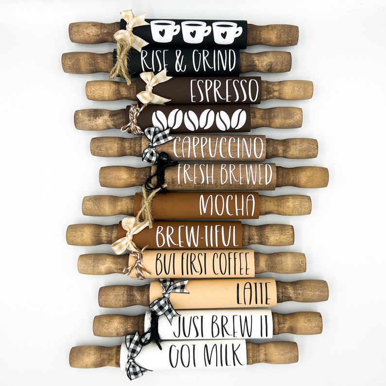 May include: A set of ten wooden rolling pins with various coffee-themed sayings painted on them. The sayings include "Rise & Grind", "Espresso", "Cappuccino", "Fresh Brewed", "Mocha", "Brew-tiful", "But First Coffee", "Latte", "Just Brew It", and "Oat Milk".