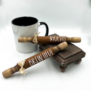 May include: Two brown wooden rolling pins with white lettering. One says "Brew-tiful" and the other says "Mocha". The rolling pins are on a small wooden stand with a white mug in the background.
