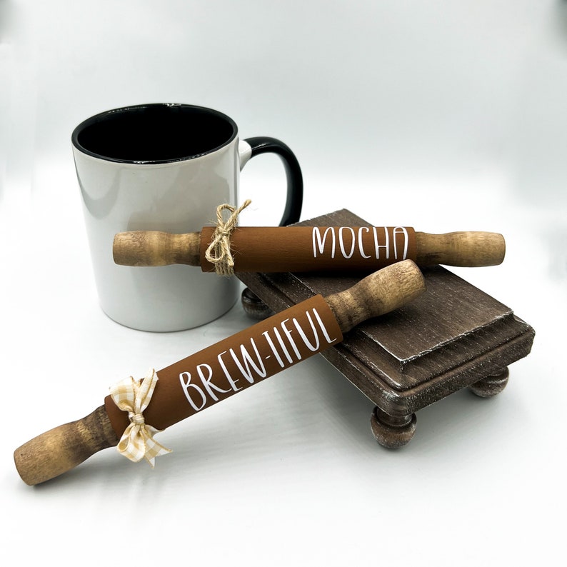 May include: Two brown wooden rolling pins with white lettering. One says "Brew-tiful" and the other says "Mocha". The rolling pins are on a small wooden stand with a white mug in the background.