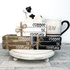 May include: Three sets of stacked wooden blocks with the text "coffee til cocktails" on each set. The blocks are in natural wood, black, and white. A white ceramic jug with the word "cream" on it and a white ceramic cow figurine are on top of the blocks.