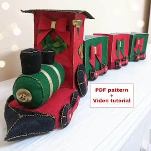 Peut inclure: Un train de Noël en feutre rouge et vert avec une locomotive noire et quatre wagons. Le train a une cloche, une couronne et des nœuds. L'image comprend le texte "PDF pattern + Video tutorial".