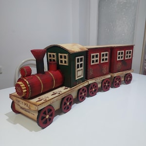 Può includere: Un treno decorativo in legno con dettagli rossi, verdi e beige. La locomotiva e i vagoni sono rossi e verdi, con ruote rosse. La base mostra le parole "NORTH POLE" e "SPECIAL DELIVERY".