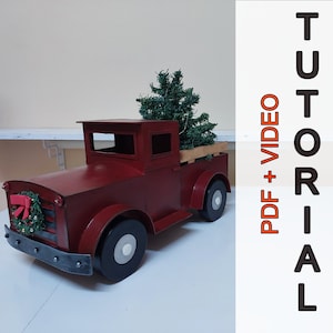 Può includere: Un camioncino giocattolo di metallo rosso con un albero di Natale artificiale nella cassone. Il camioncino ha un design a strisce bianche e nere sul lato e un fiocco rosso sul davanti.
