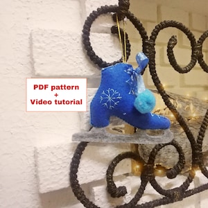 Könnte beinhalten: Blauer Filz-Schlittschuh-Anhänger mit einem weißen Schneeflocken-Design und einem blauen Bommel. Der Anhänger hängt an einem schwarzen Metallhaken. Der Text "PDF pattern + Video tutorial" ist im Bild sichtbar.