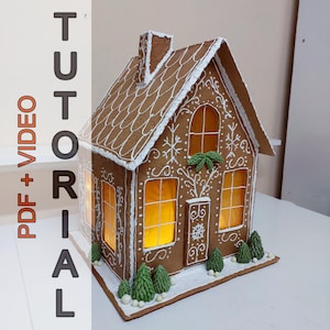 Può includere: Una casetta di pan di zenzero con glassa bianca e un interno illuminato. La casa ha un tetto marrone, modanature bianche e un piccolo camino. Le finestre sono decorate con fiocchi di neve e la porta ha un motivo a fiocco di neve. La casa è circondata da piccoli alberi verdi e neve bianca.