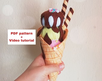 May include: A felt ice cream cone with three scoops of ice cream: chocolate, vanilla, and strawberry. The cone is decorated with sprinkles and two chocolate sticks. The text "PDF pattern + Video tutorial" is on a white rectangle.