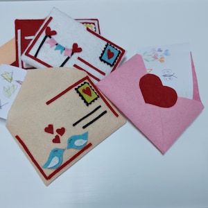 Pode incluir: Um conjunto de envelopes de feltro com cora&ccedil;&otilde;es vermelhos, p&aacute;ssaros azuis e um selo. Os envelopes s&atilde;o decorados com uma faixa vermelha, branca e azul.