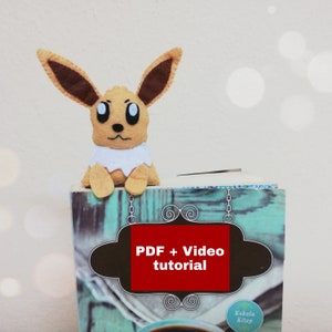 May include: A brown and tan felt Eevee plush toy bookmark with white fur around its neck. The bookmark is sitting on top of a book with a teal cover. The book has a brown sign with the text "PDF + Video tutorial" in red letters.