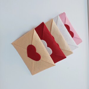 Pode incluir: Cinco envelopes de feltro em v&aacute;rias cores, incluindo bege, branco, vermelho e rosa, com um cora&ccedil;&atilde;o vermelho recortado em cada um.