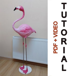 Pode incluir: Uma escultura de flamingo rosa de papel maché em pé sobre uma base branca. O flamingo está em um piso de madeira na frente de um radiador branco. O texto "PDF + VIDEO TUTORIAL" está no canto superior direito da imagem.