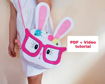 Puede incluir: Un bolso de fieltro blanco con forma de conejo con orejas rosas, gafas rosas y flores de colores. El bolso tiene una correa rosa y lo lleva puesto un niño. PDF + Tutorial en vídeo.