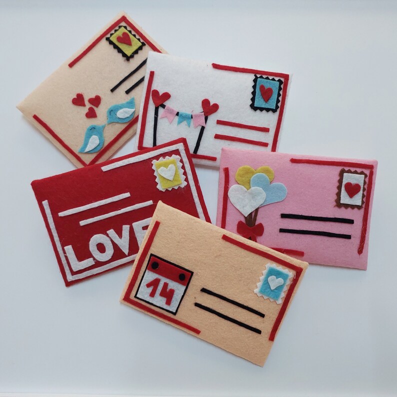 Pode incluir: Cinco envelopes de feltro com diferentes designs. Os envelopes s&atilde;o decorados com cora&ccedil;&otilde;es, p&aacute;ssaros, bal&otilde;es, um calend&aacute;rio com o n&uacute;mero 14 e a palavra "LOVE".