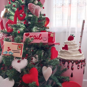 Pode incluir: Uma caixa de correio rosa e branca com a palavra "mail" nela, cercada por uma &aacute;rvore de Natal decorada com cora&ccedil;&otilde;es vermelhos e um milkshake rosa e branco com um morango no topo.