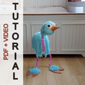 May include: A quirky, handmade bird sculpture. The bird has a light blue yarn body, a pink yarn neck, and brown feet. It features a large orange beak and big, expressive eyes. The text "PDF + VIDEO TUTORIAL" is visible on the left side.